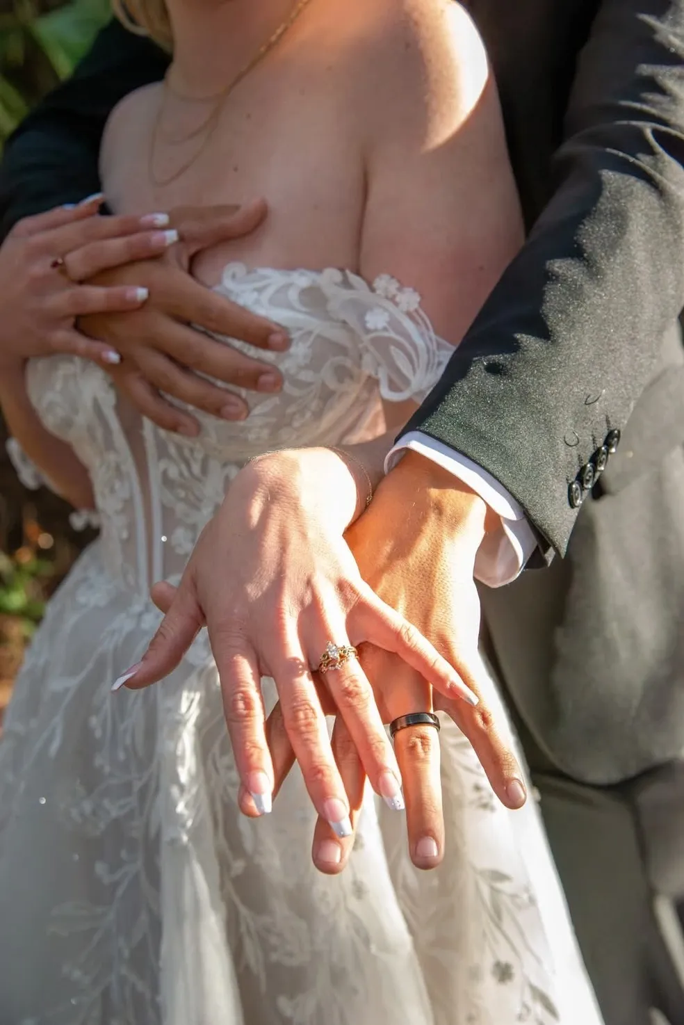 A couple in wedding attire embraces, their hands joined in the foreground to showcase their wedding bands.