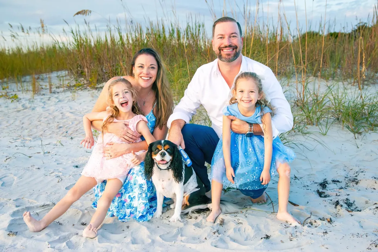 A family with a dog poses on a sandy beach at sunset, with tall grass in the background.
