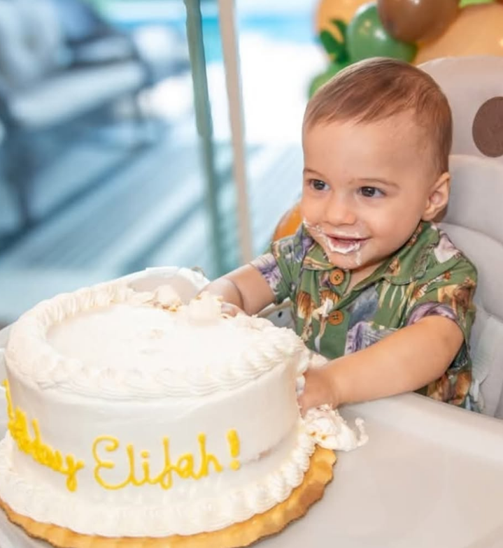 A toddler in a patterned shirt with cake on their face reaches into a white birthday cake labeled 