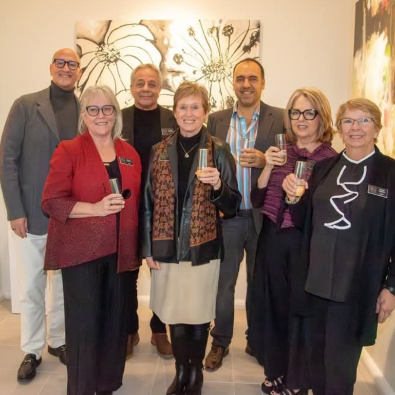Seven people smiling and holding drinks pose in an art gallery, standing in front of large black-and-white floral artwork.