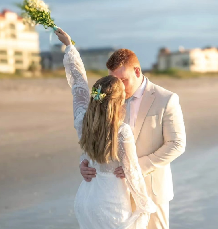 A couple in wedding attire stands on a beach, the partner with their arm raised high holding a bouquet toward the sky.