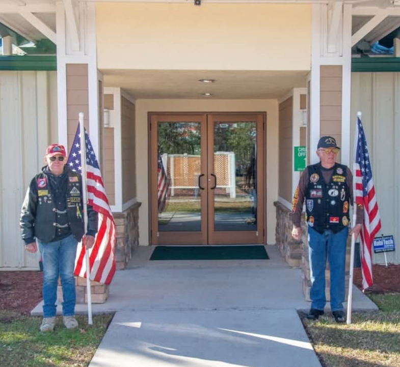 Two veterans in vests stand outside a building entrance, each holding an American flag on a pole.