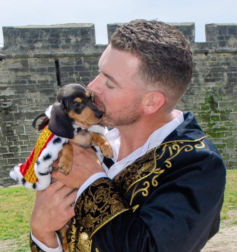 A person in an ornate, gold-trimmed black coat kisses a puppy wearing a tiny red and white cape in front of a stone wall.