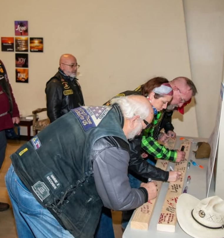 People wearing leather vests sign a wooden board at a table in a room with wall art.