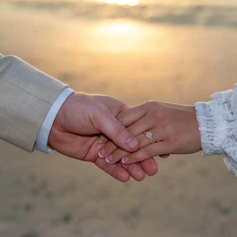A close-up of two people holding hands at sunset on a beach, with a prominent diamond ring visible on one finger.