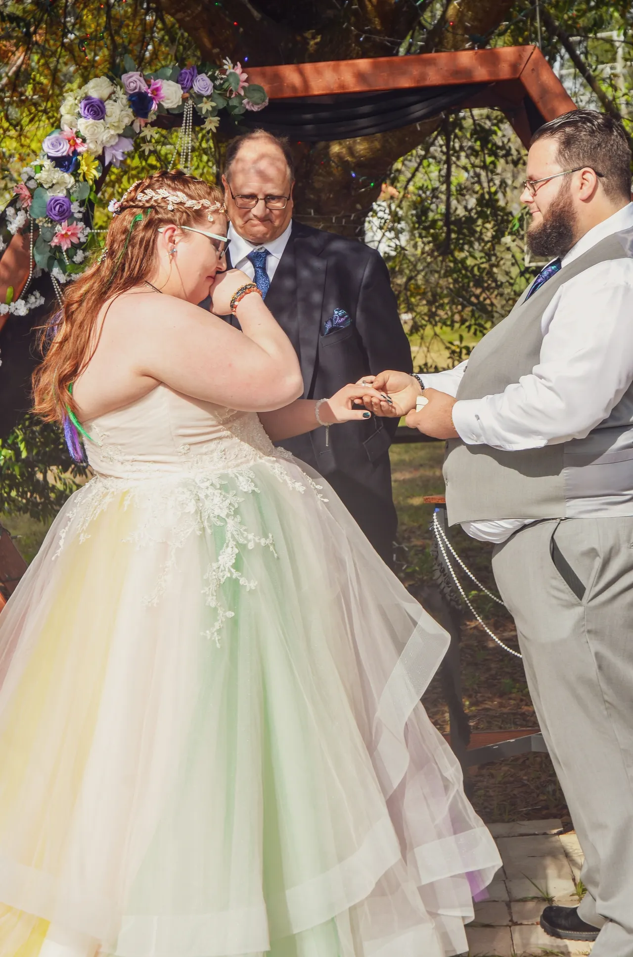 A person in a pastel-colored gown emotionally exchanges rings with a partner in a grey vest during an outdoor wedding.