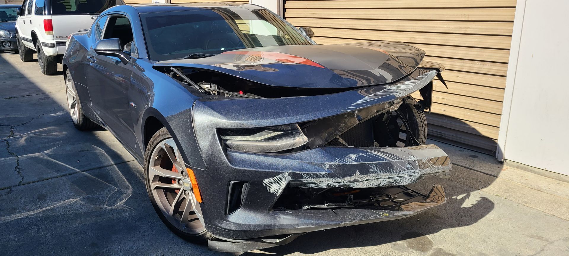 A black car with a damaged front end is parked on the side of the road.