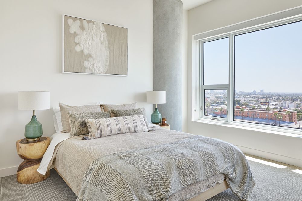 Bright bedroom with a bed, bedside lamps, and a large window overlooking a city skyline