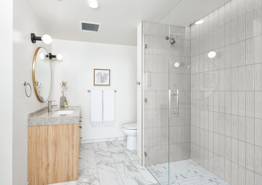 Bright white bathroom with marble floor, wood vanity, round mirror, toilet, and glass shower enclosure.