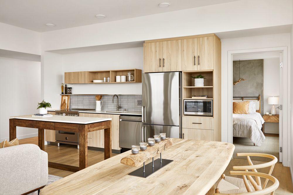 Bright open kitchen and dining area with light wood furniture, stainless steel appliances, and a bedroom visible through a doorway