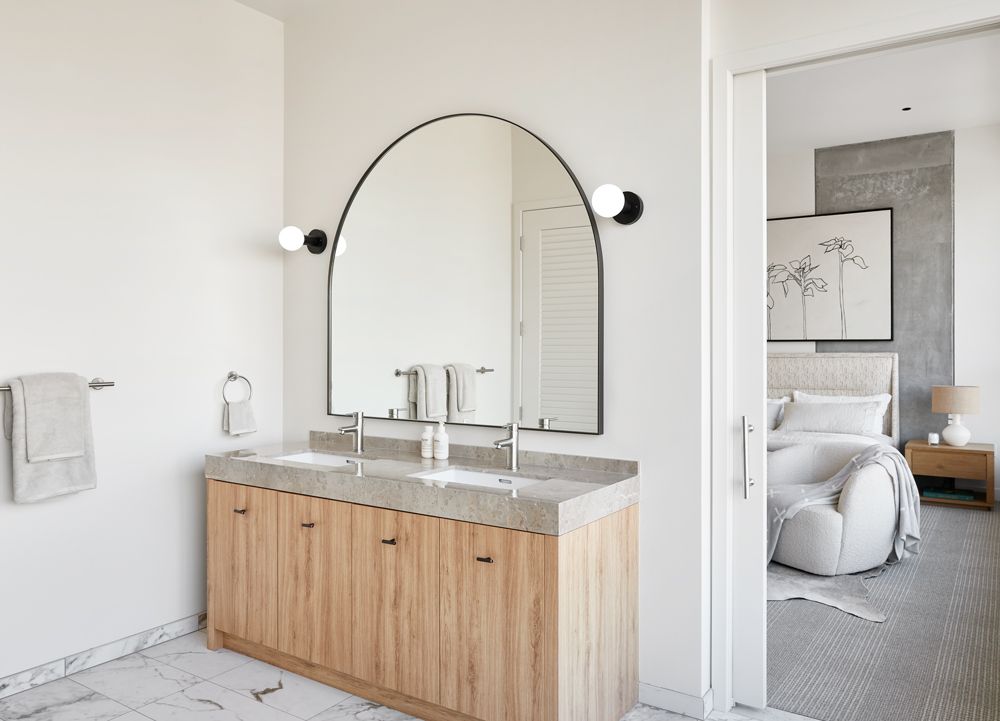 Modern bathroom vanity with arched mirror, wall sconces, and open doorway to a bedroom.
