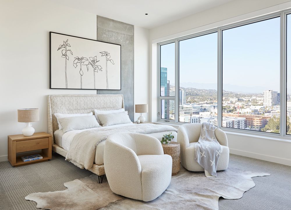 Bright modern bedroom with white bed, lounge chair, and large windows overlooking a city skyline