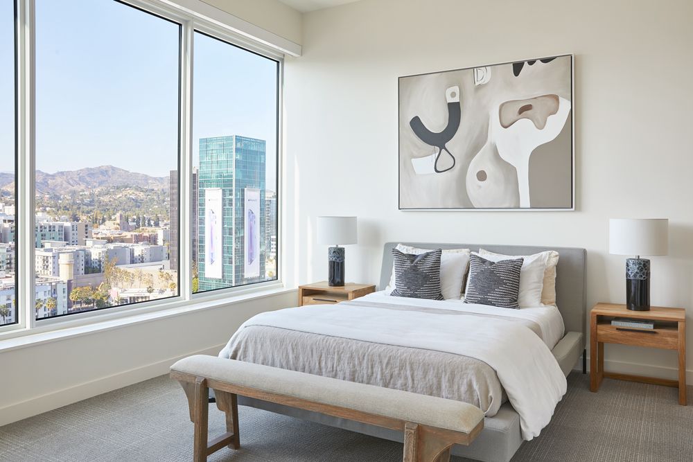 Bright modern bedroom with large windows, city view, and a neatly made bed with abstract wall art