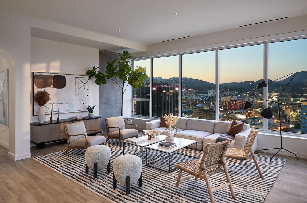 Modern living room with beige sectional, striped rug, woven chairs, and city view through floor-to-ceiling windows