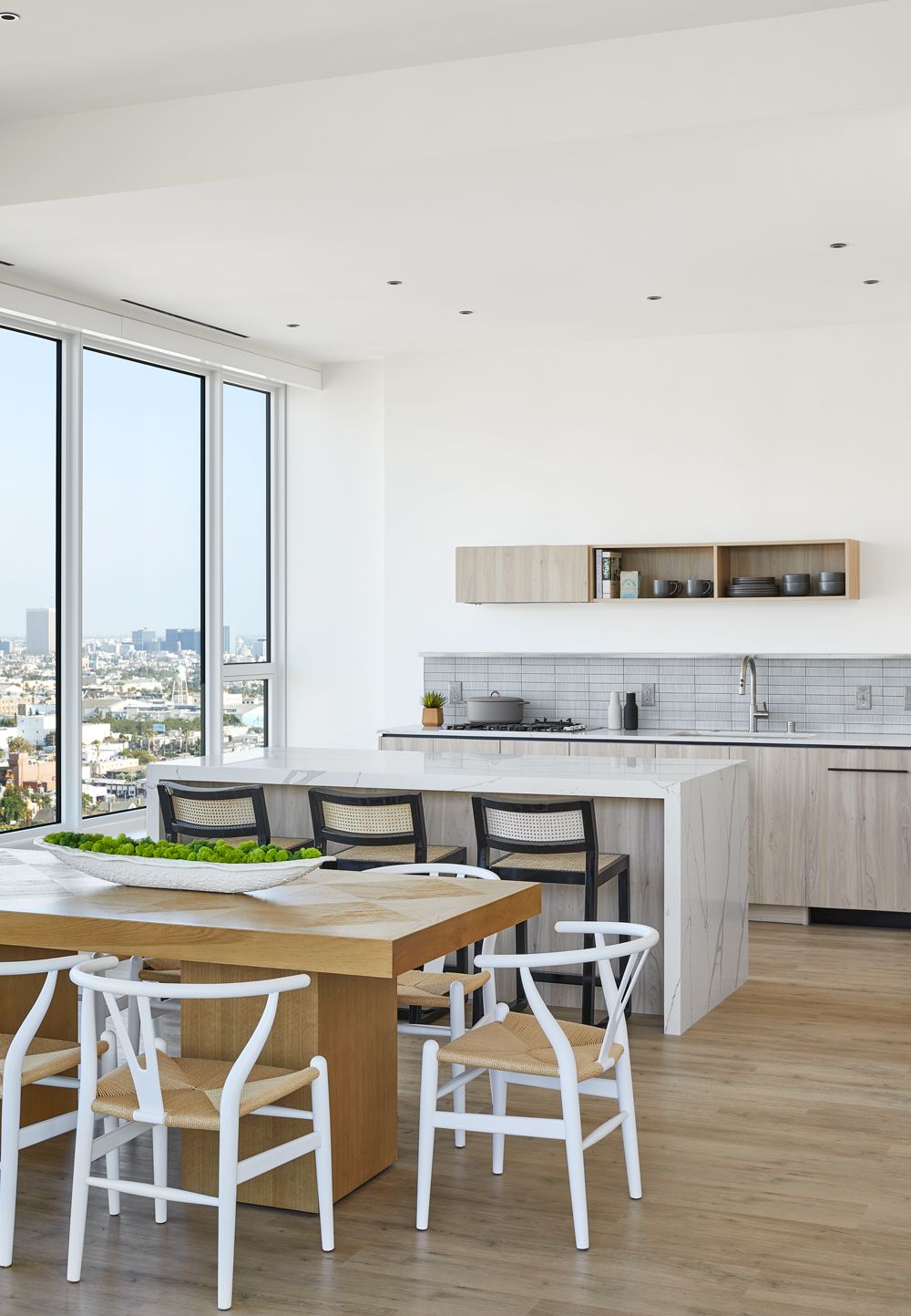 Bright modern kitchen with wooden dining table, white chairs, island, and large windows overlooking a city.