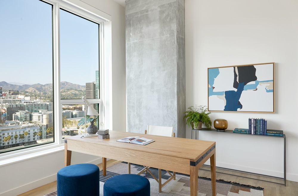 Bright modern office with a wooden desk, blue stools, wall art, and a city view through large windows