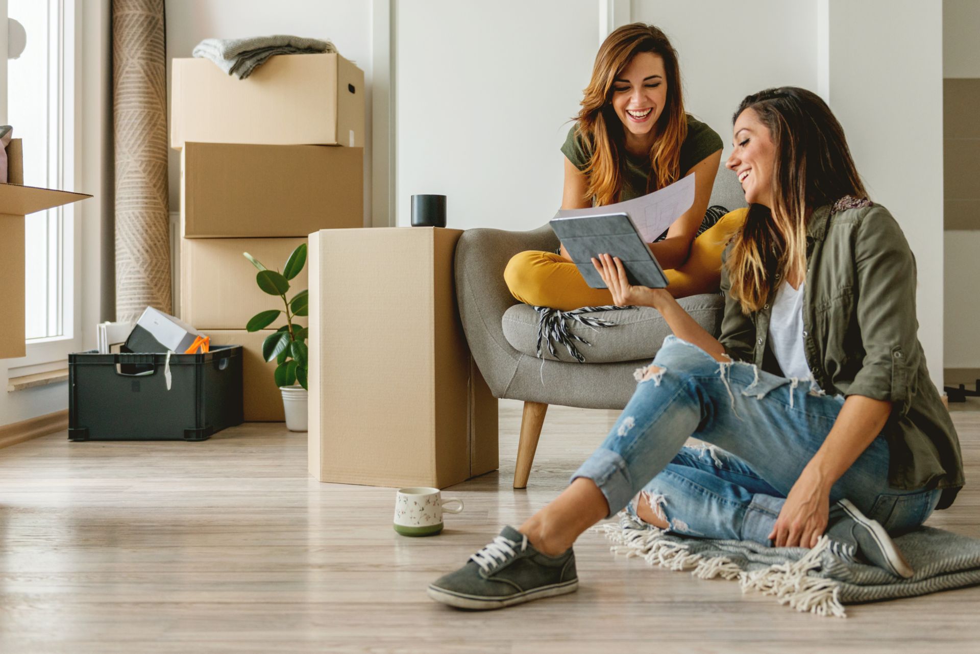 Two people sitting on the floor in a bright room, smiling at a tablet during unpacking after a move