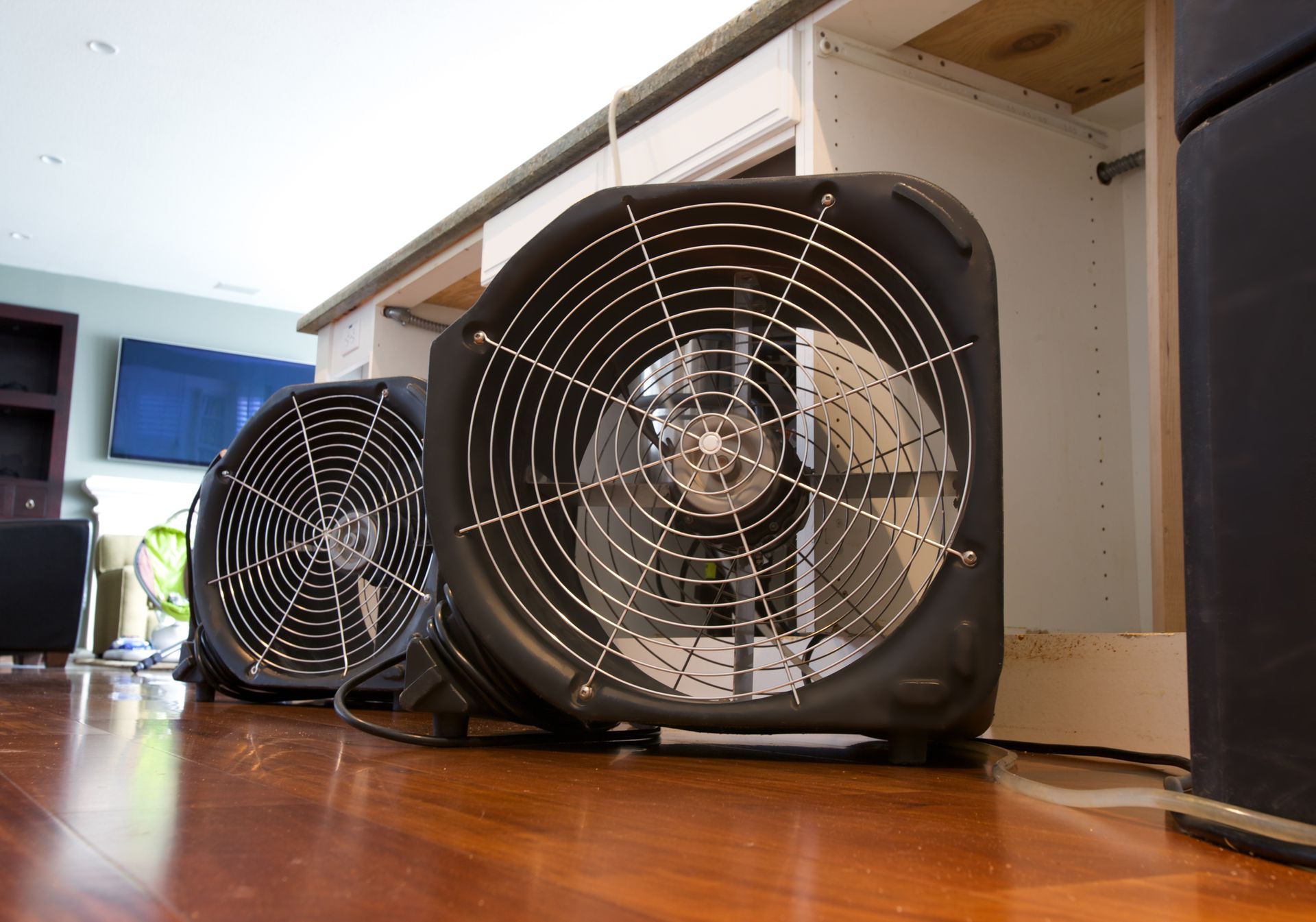 Two black industrial fans sit on a hardwood floor, positioned to dry a set of kitchen cabinets.