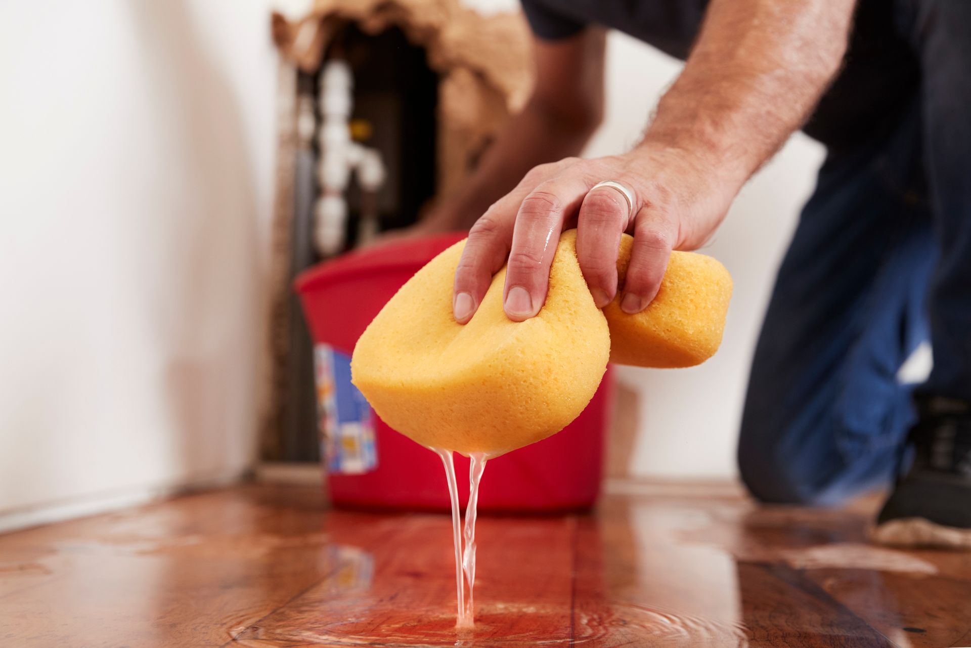 A person using a yellow sponge to soak up water from a wooden floor near an exposed wall and a red plastic container.