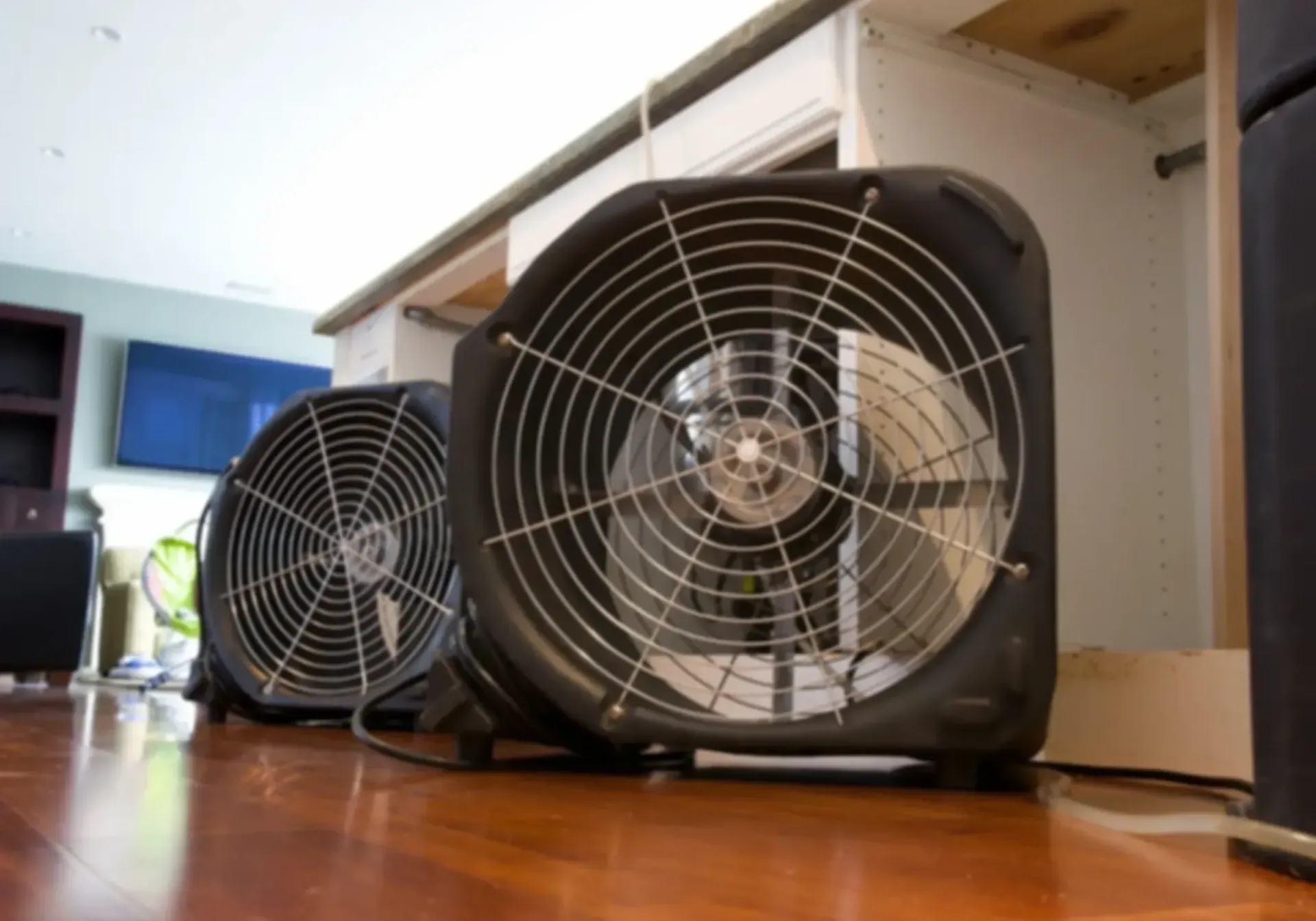 Two industrial floor fans placed on a wood floor in a home for drying or ventilation.