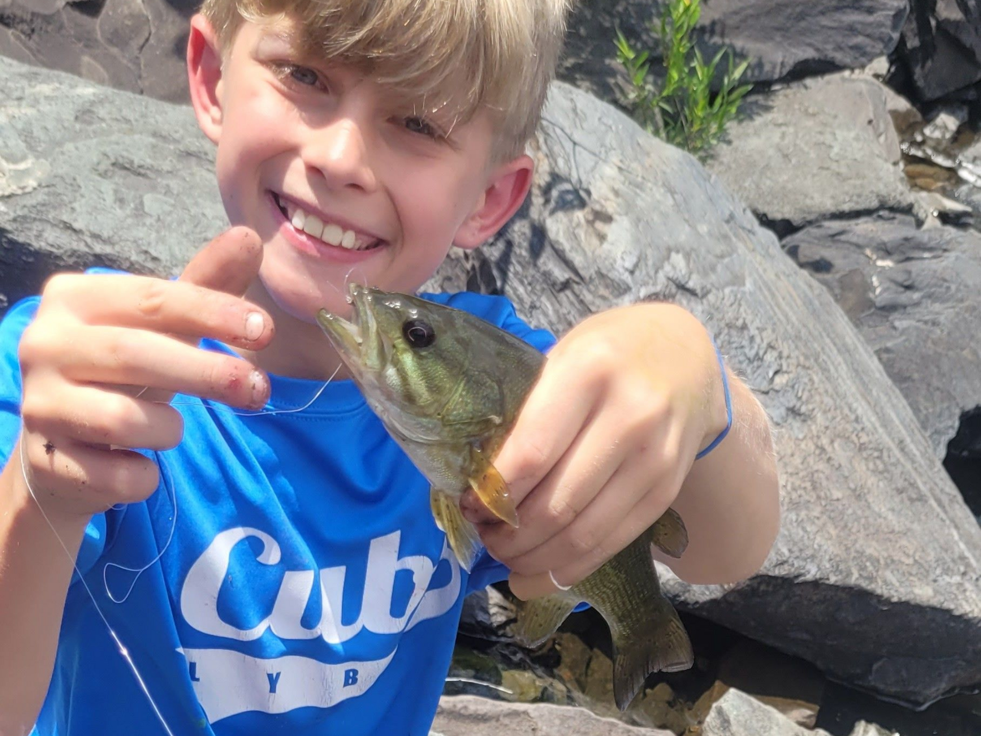 Boy holding a fish and smiling outdoors on rocks by the water