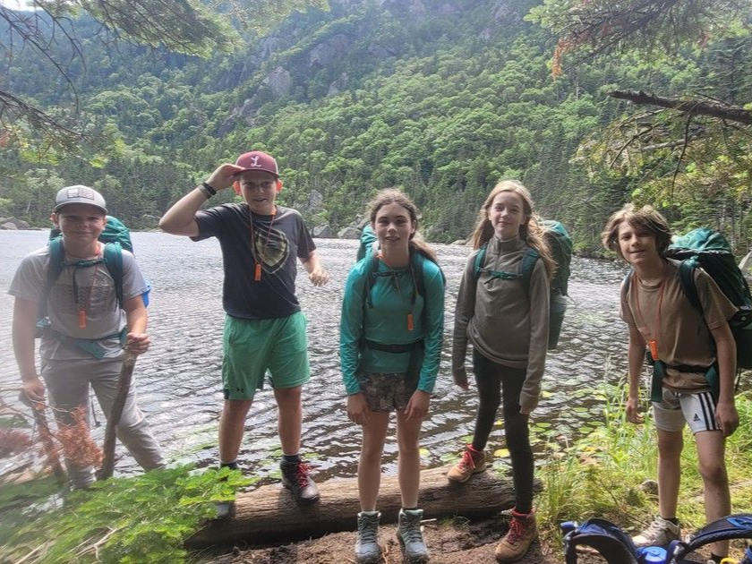 Five hikers standing on a log by a mountain stream in a lush forest