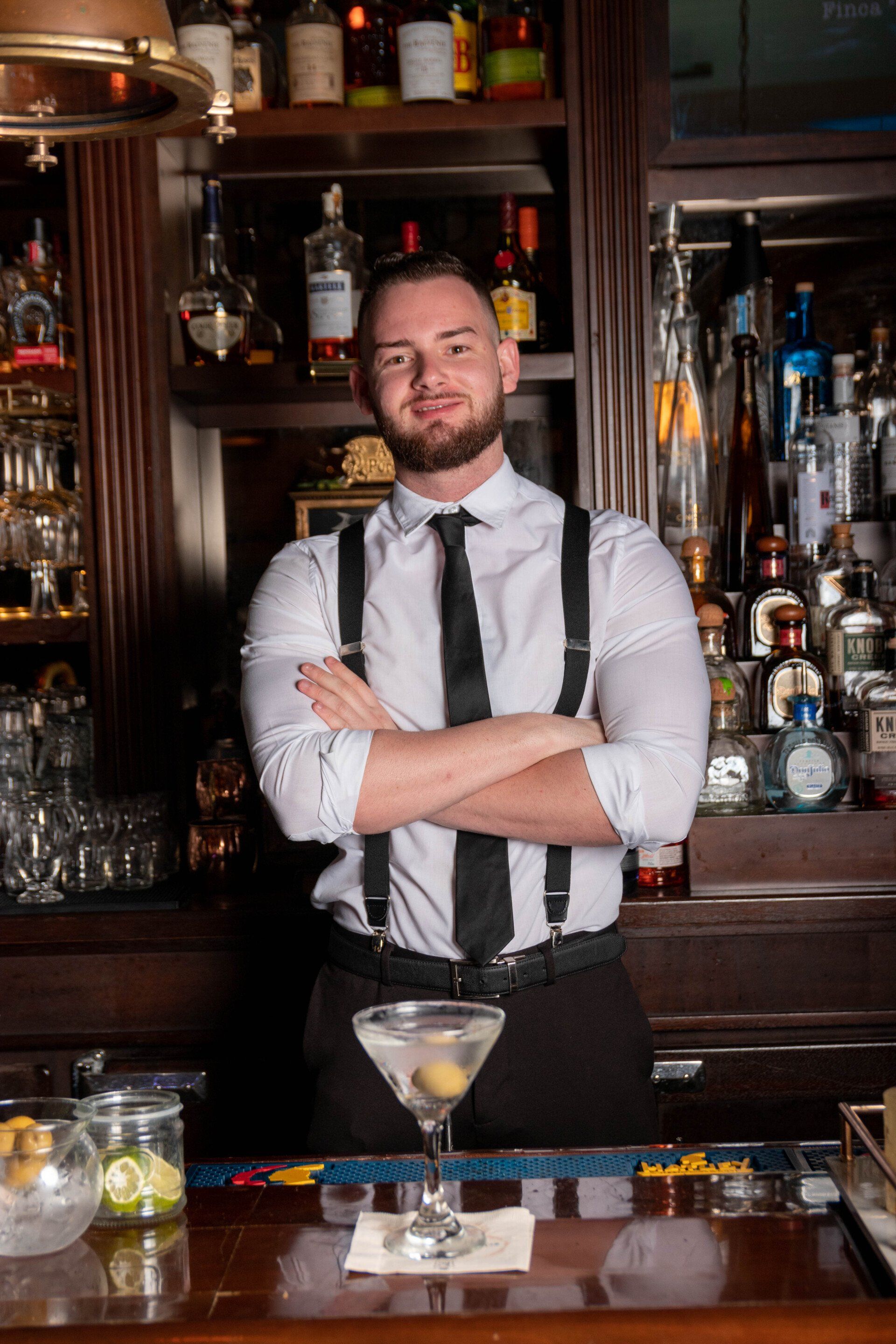 A bartender is standing behind a bar with his arms crossed next to a martini glass.