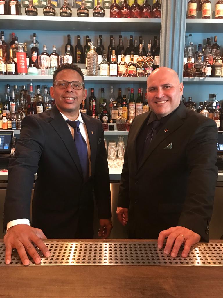 Two men in suits and ties are standing behind a bar.