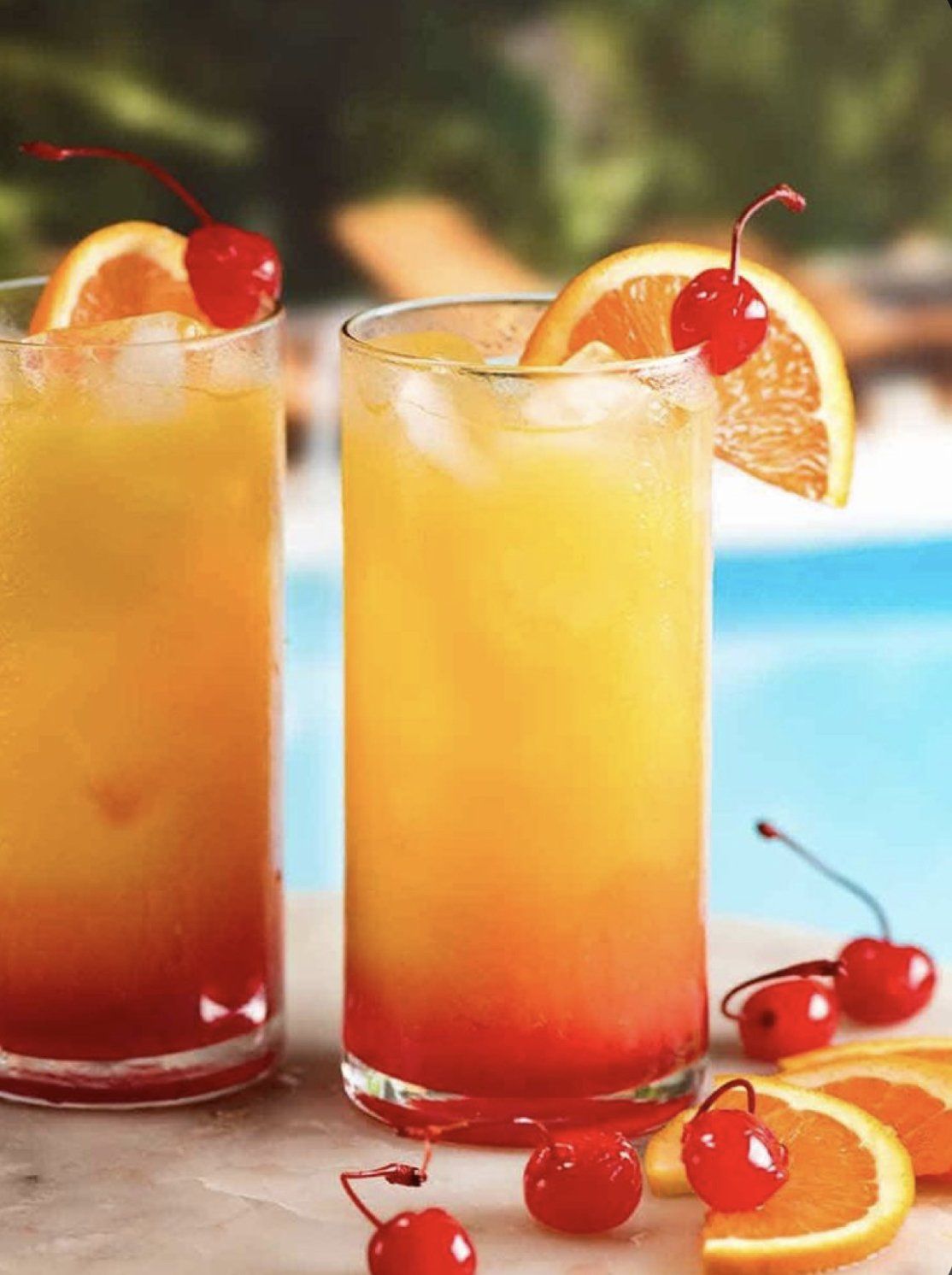Two glasses of tequila sunrise cocktails with cherries and oranges on a table.