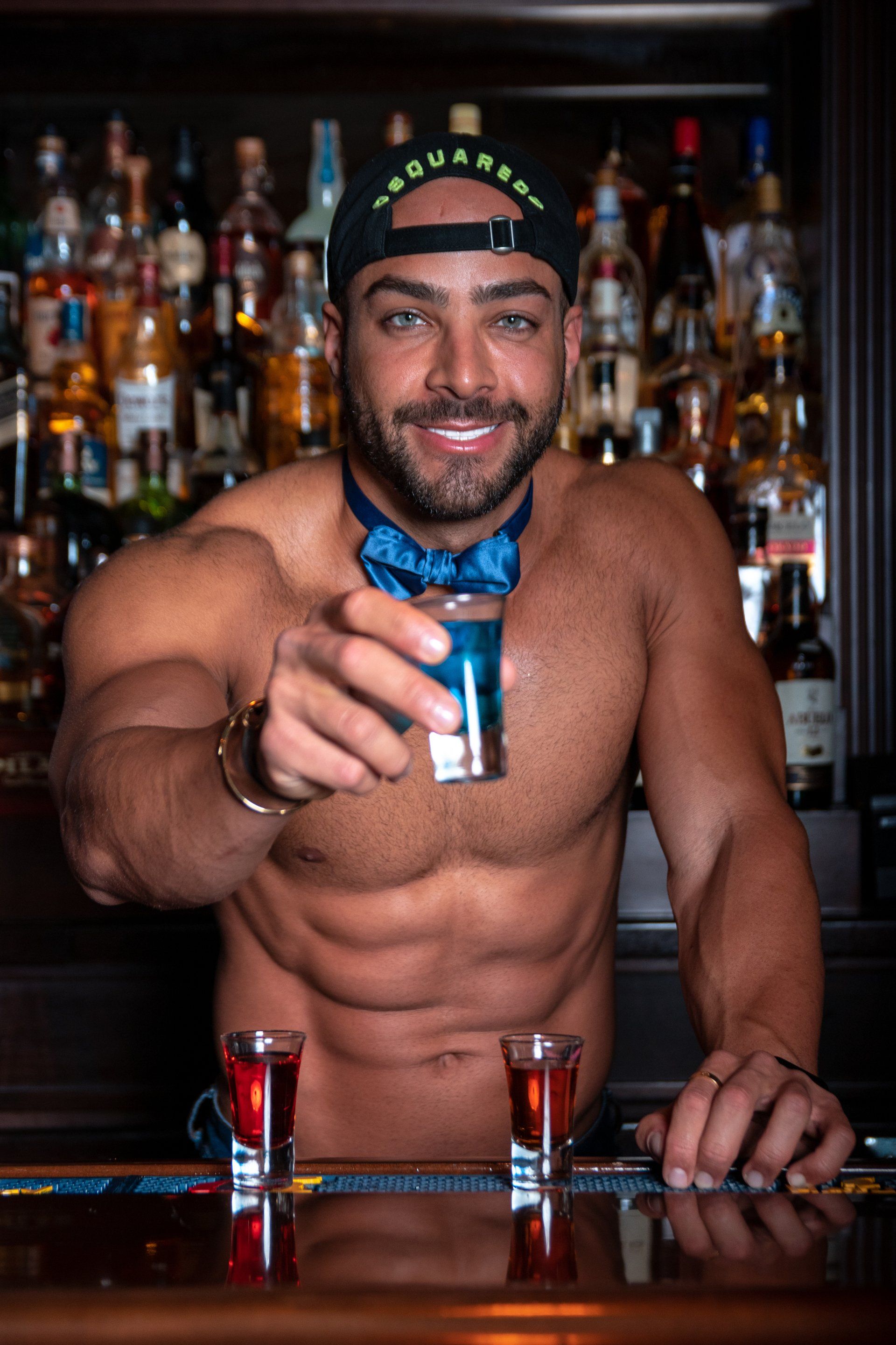 A shirtless man is standing at a bar holding a shot glass.