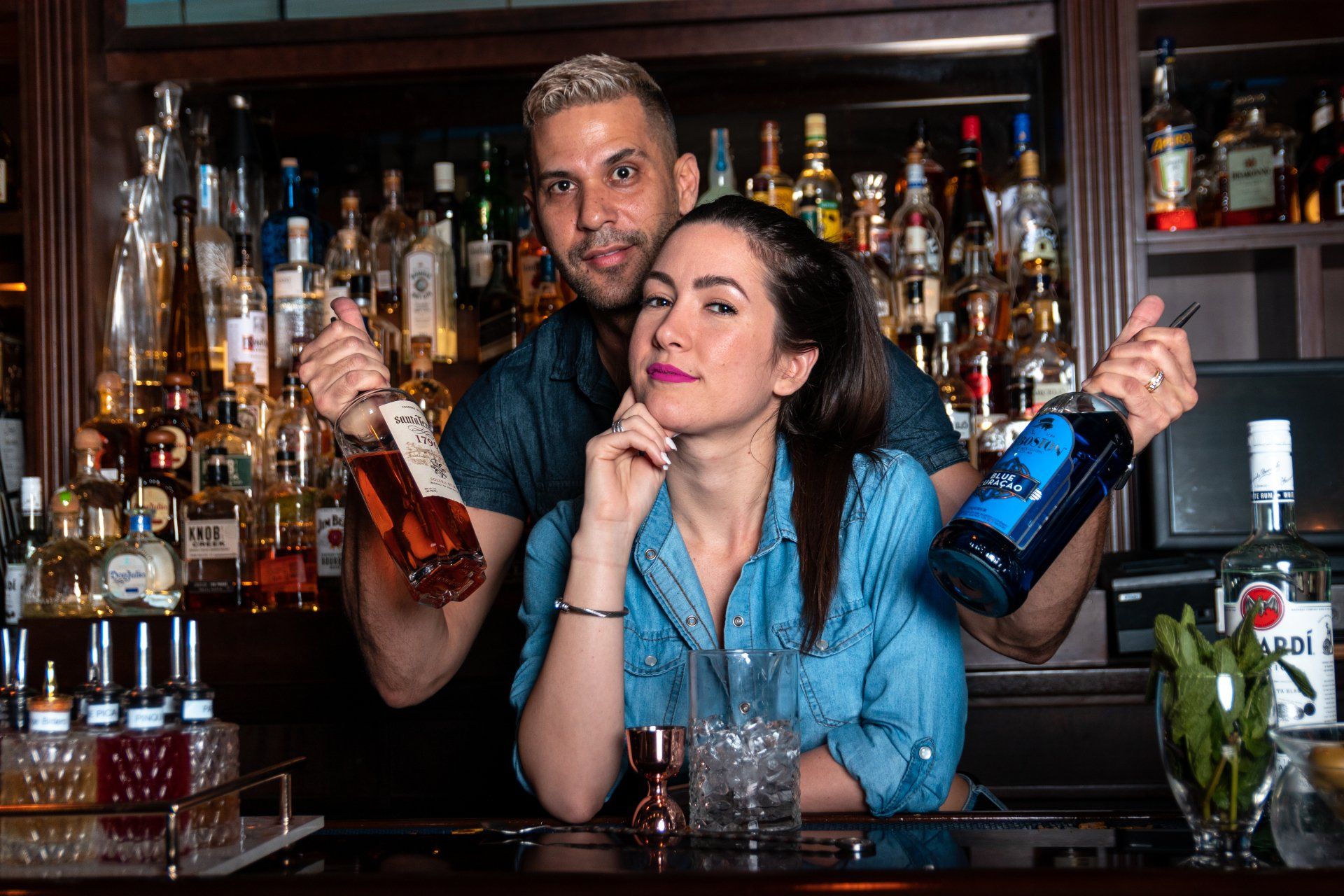 A man and a woman are standing behind a bar holding bottles of alcohol.
