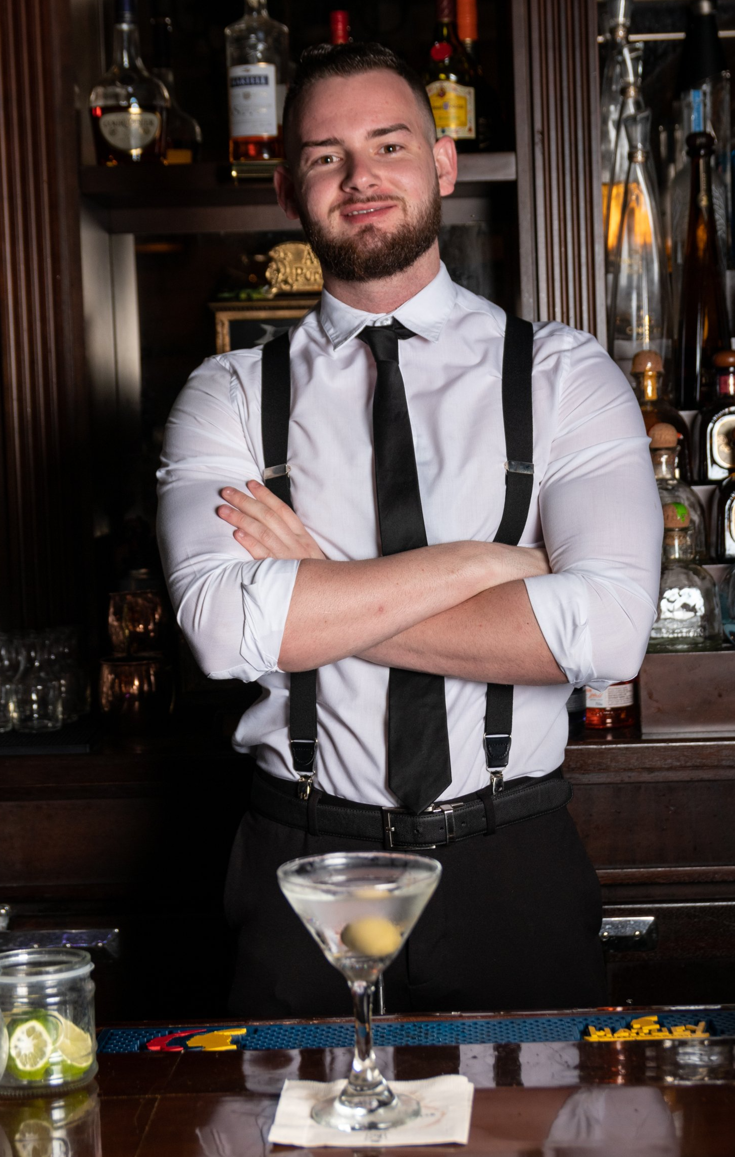 A man in a white shirt and black tie is standing behind a bar with his arms crossed.