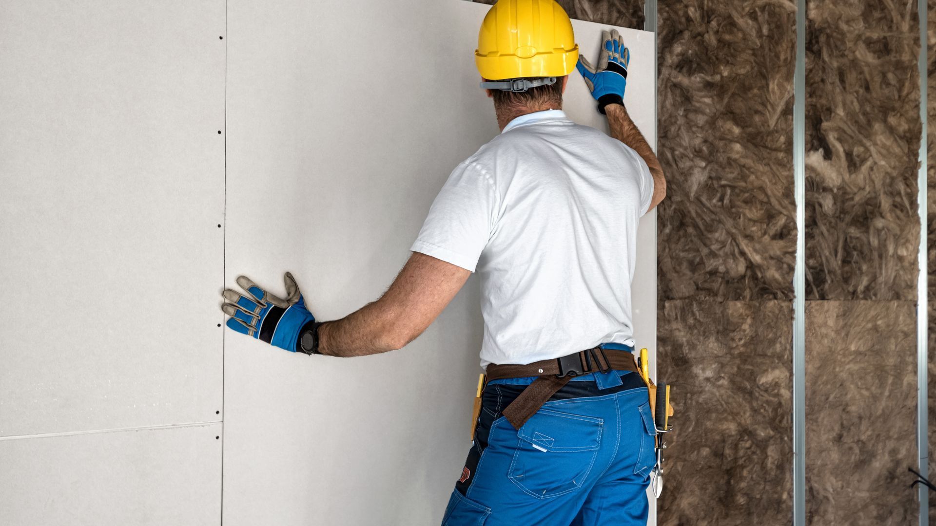 Worker in yellow hard hat installing white wall panels beside exposed insulation.