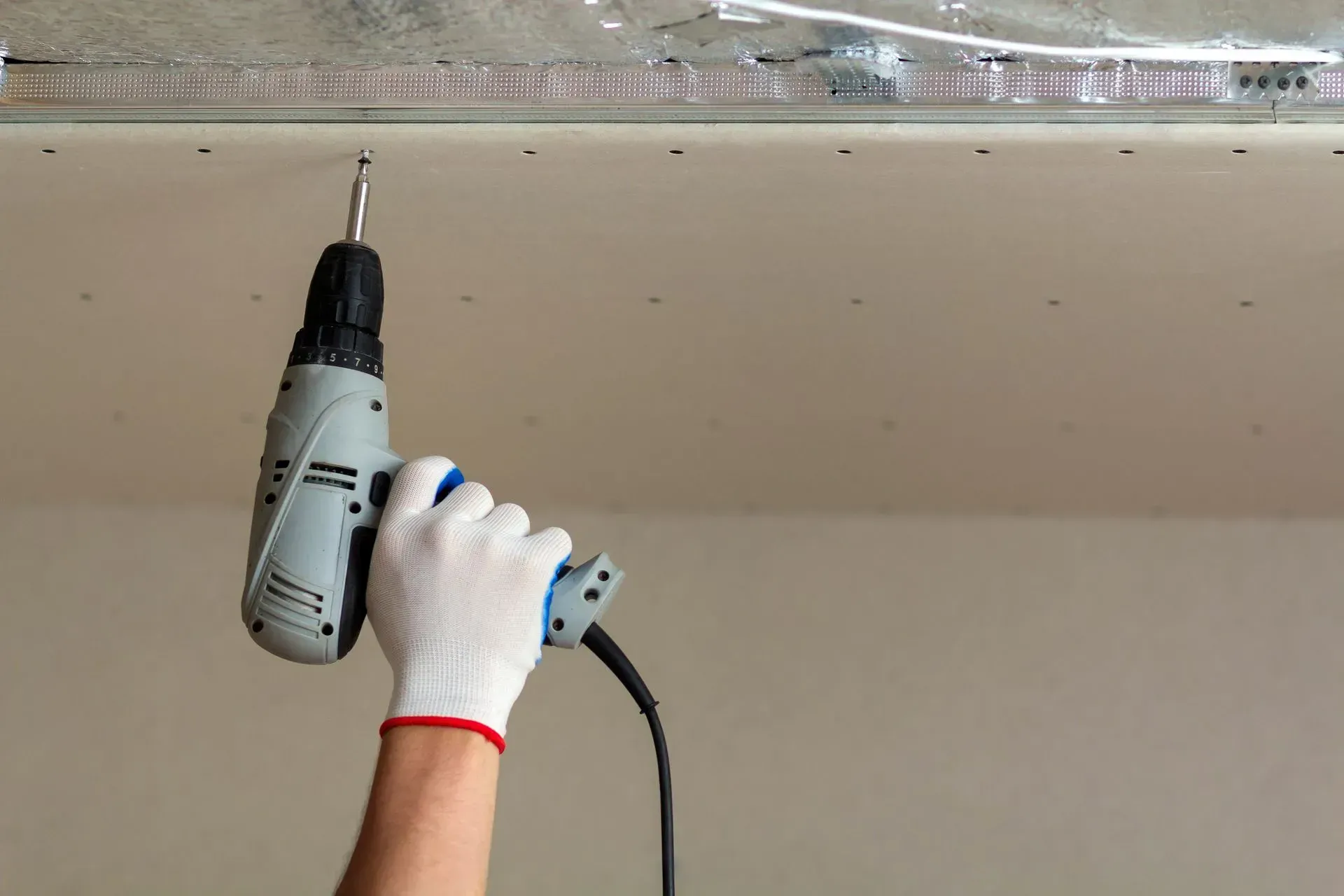 Gloved hand using a power drill on a ceiling seam with clear tape.