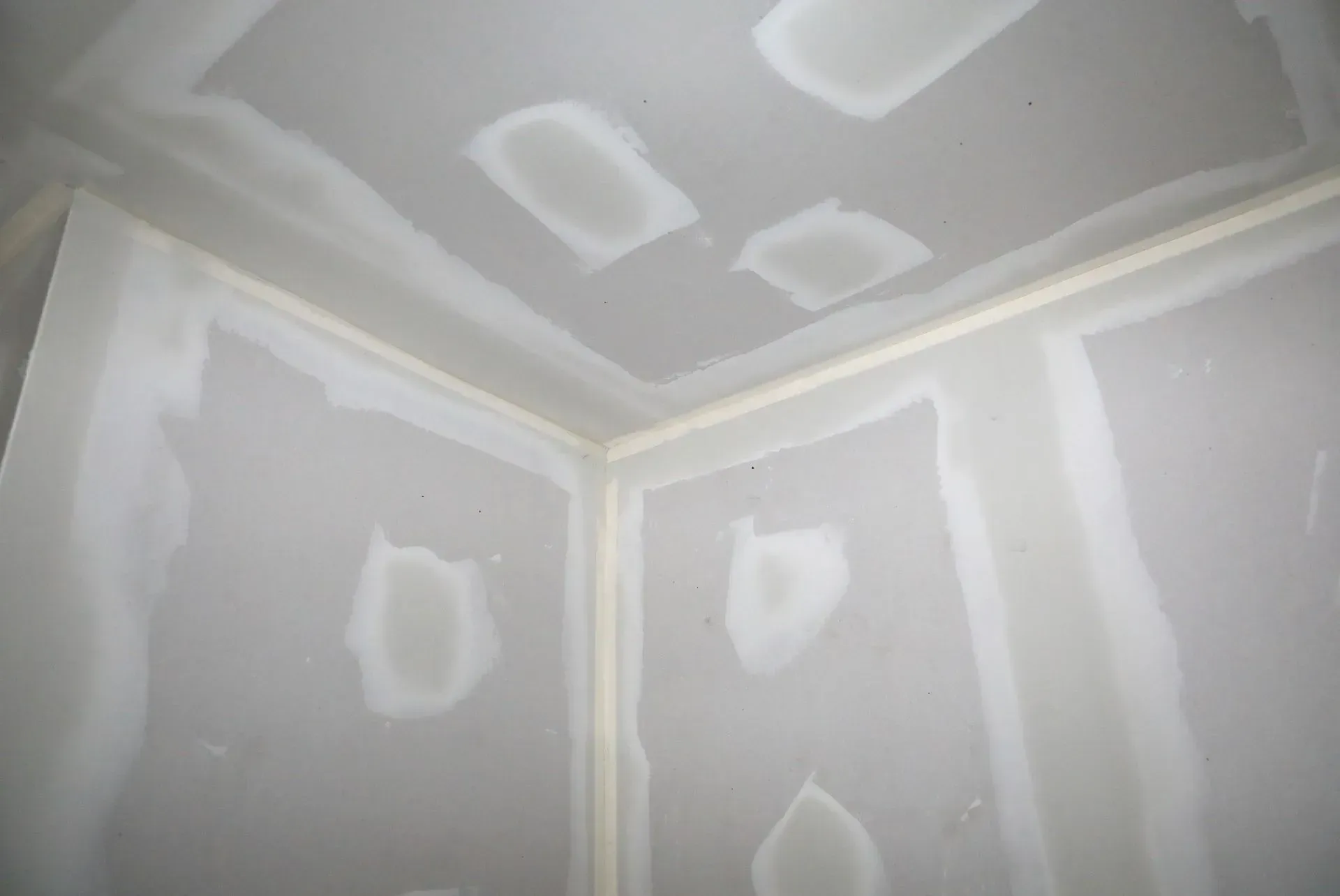 Unfinished drywall room corner with taped seams and white patch marks on ceiling and walls