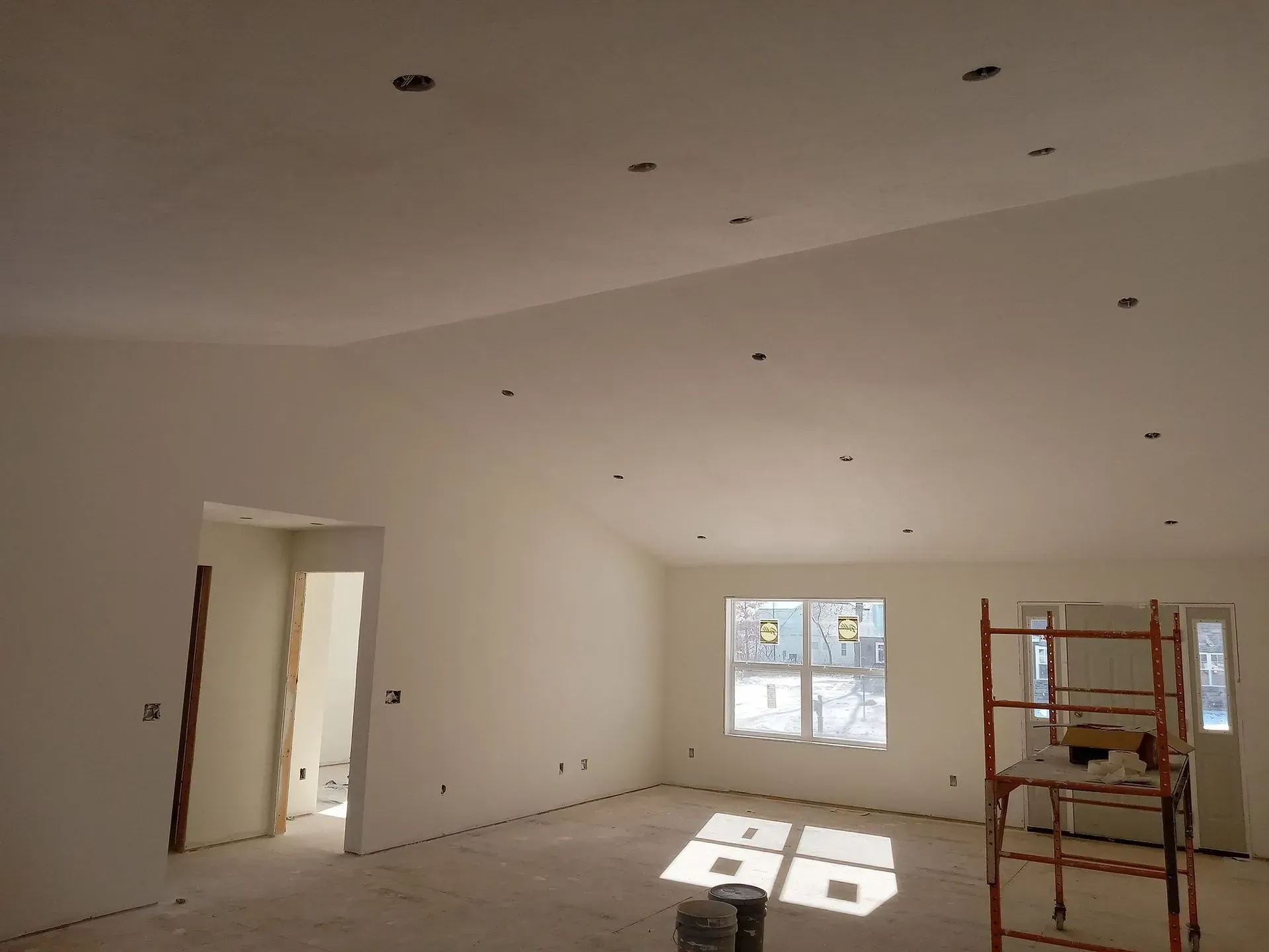 Empty white room under renovation with drywall, ceiling lights, and scaffold by a window