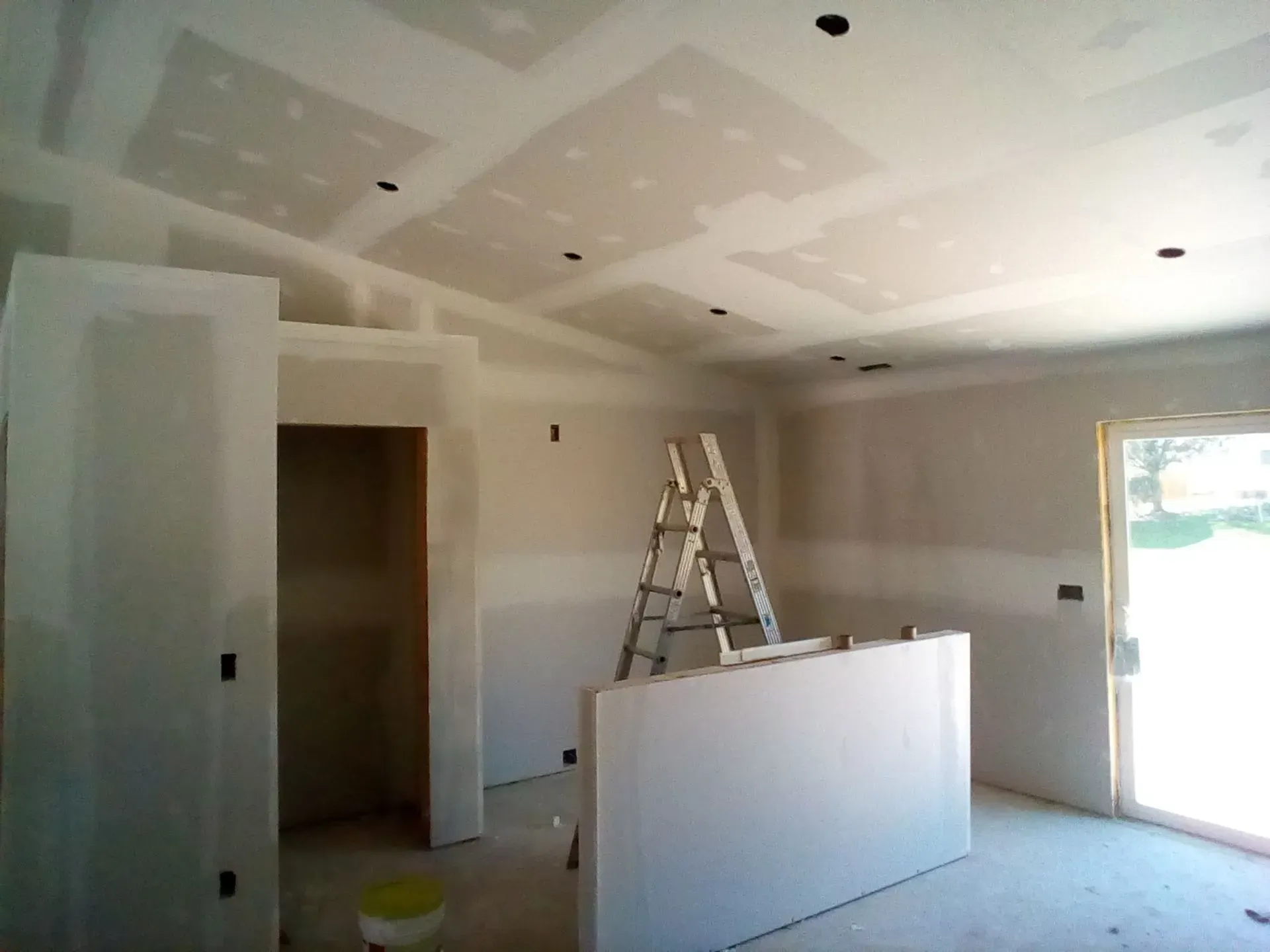 Interior under renovation with drywall, ladder, and an open door to outside.