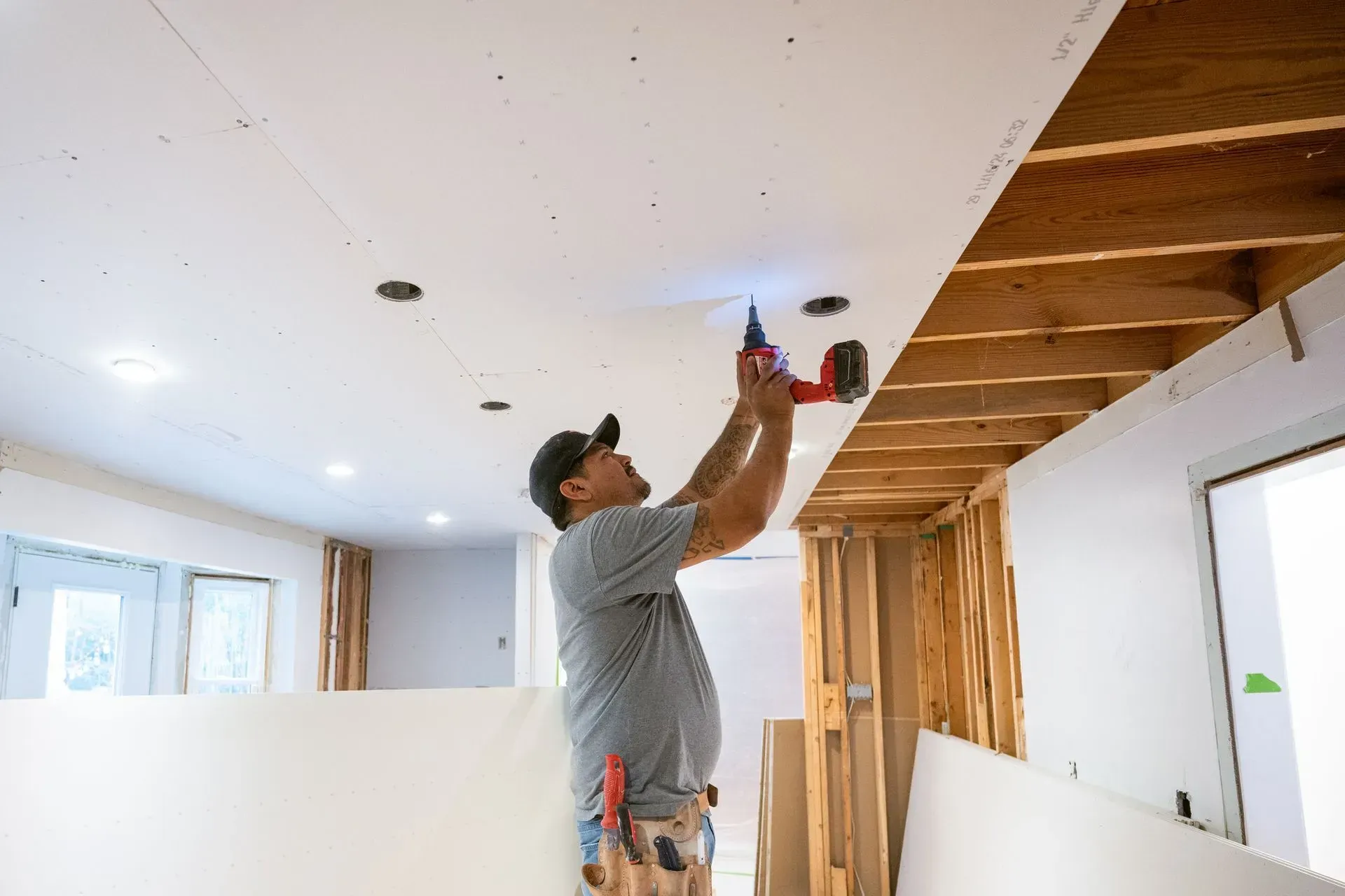 Worker installing ceiling drywall with a power drill in an unfinished room