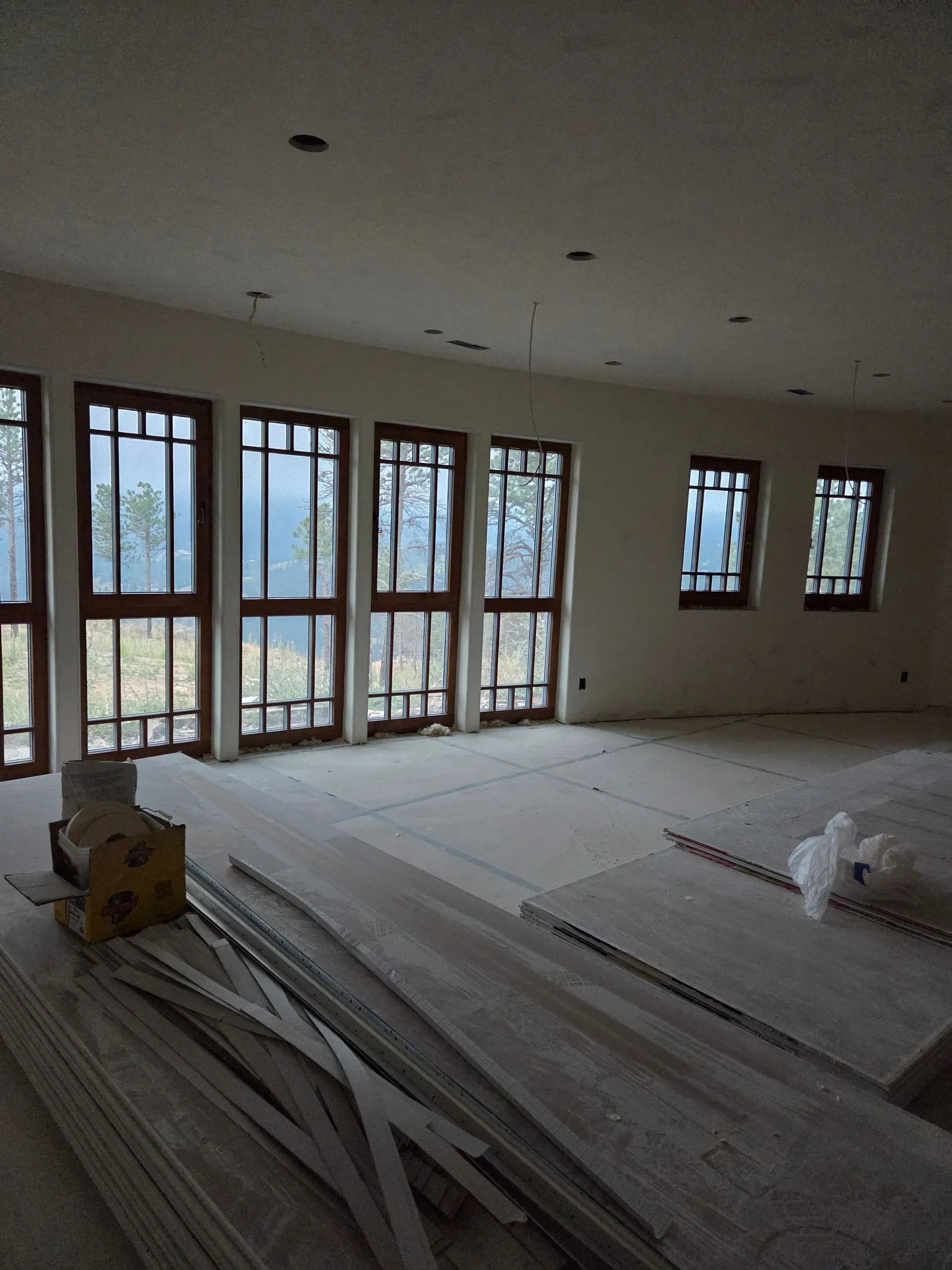 Empty room under construction with large windows, bare floors, and scattered building materials.