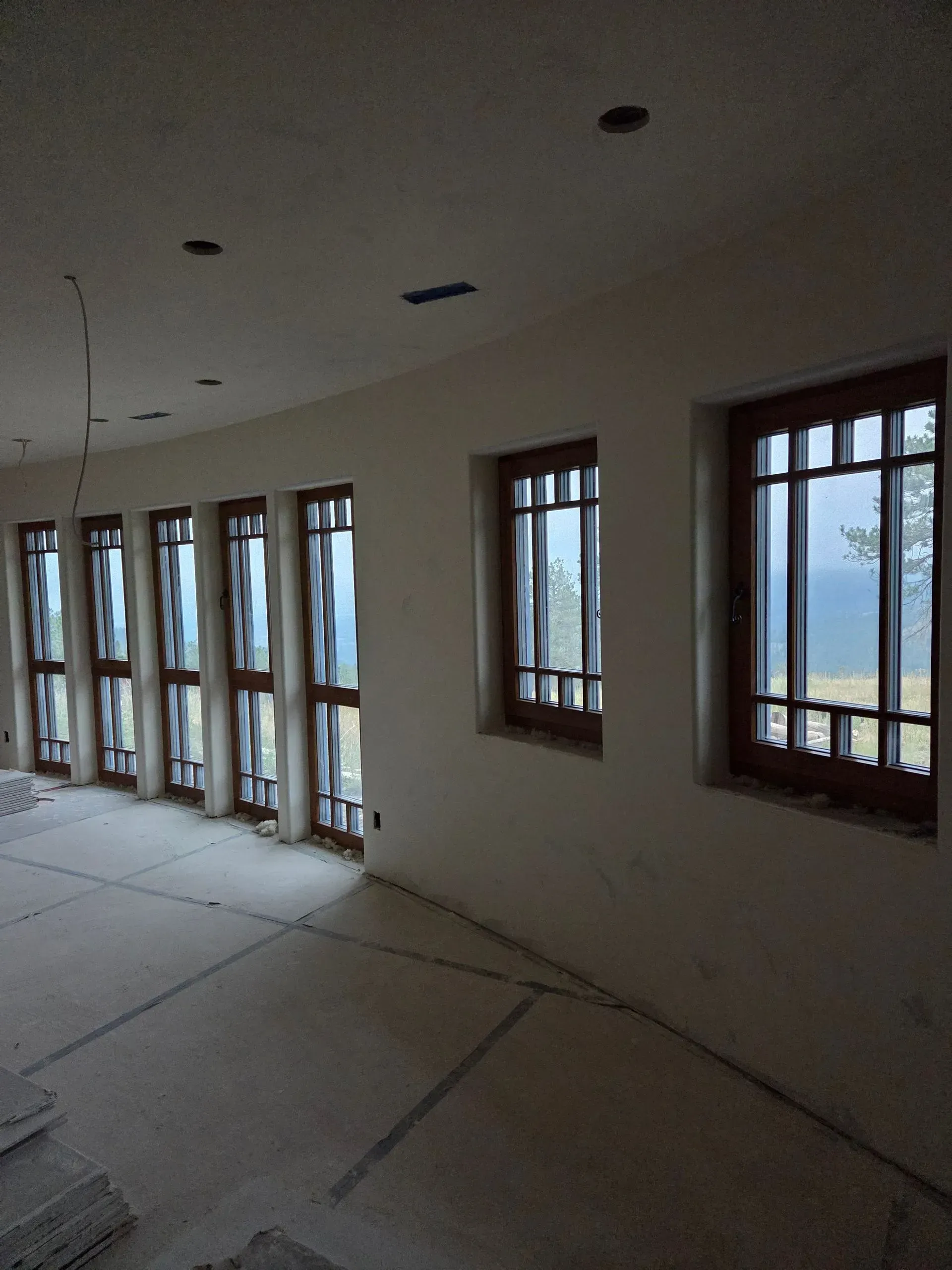 Empty room with unfinished floor and walls, lined with windows overlooking a landscape.