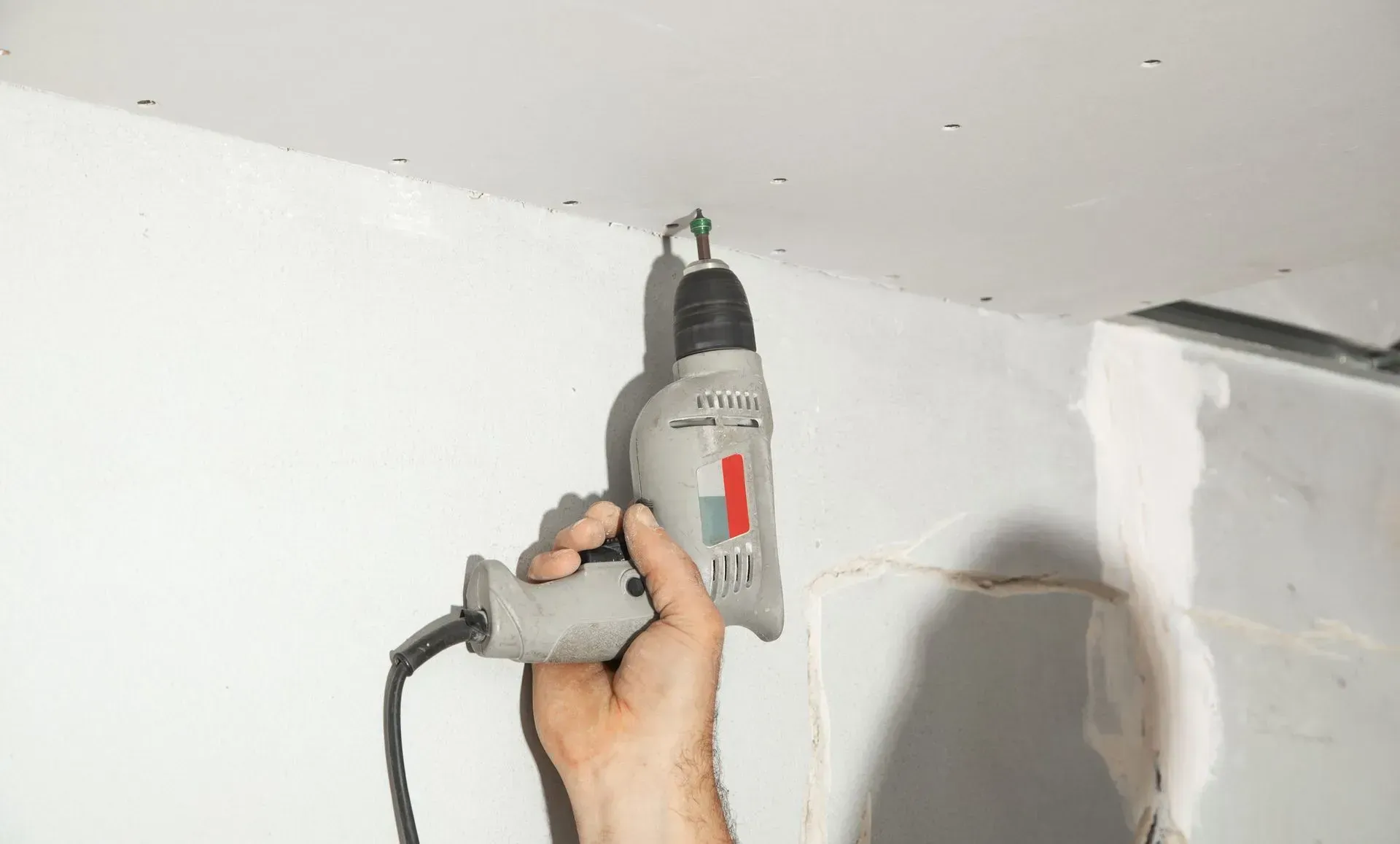 Hand holding a gray electric drill against a white ceiling, drilling into a wall corner.