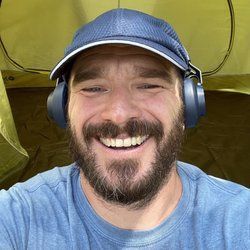 Smiling bearded man in a blue cap and headphones, taking a selfie in a yellow tent