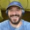 Smiling bearded man in a blue cap and headphones, taking a selfie in a yellow tent