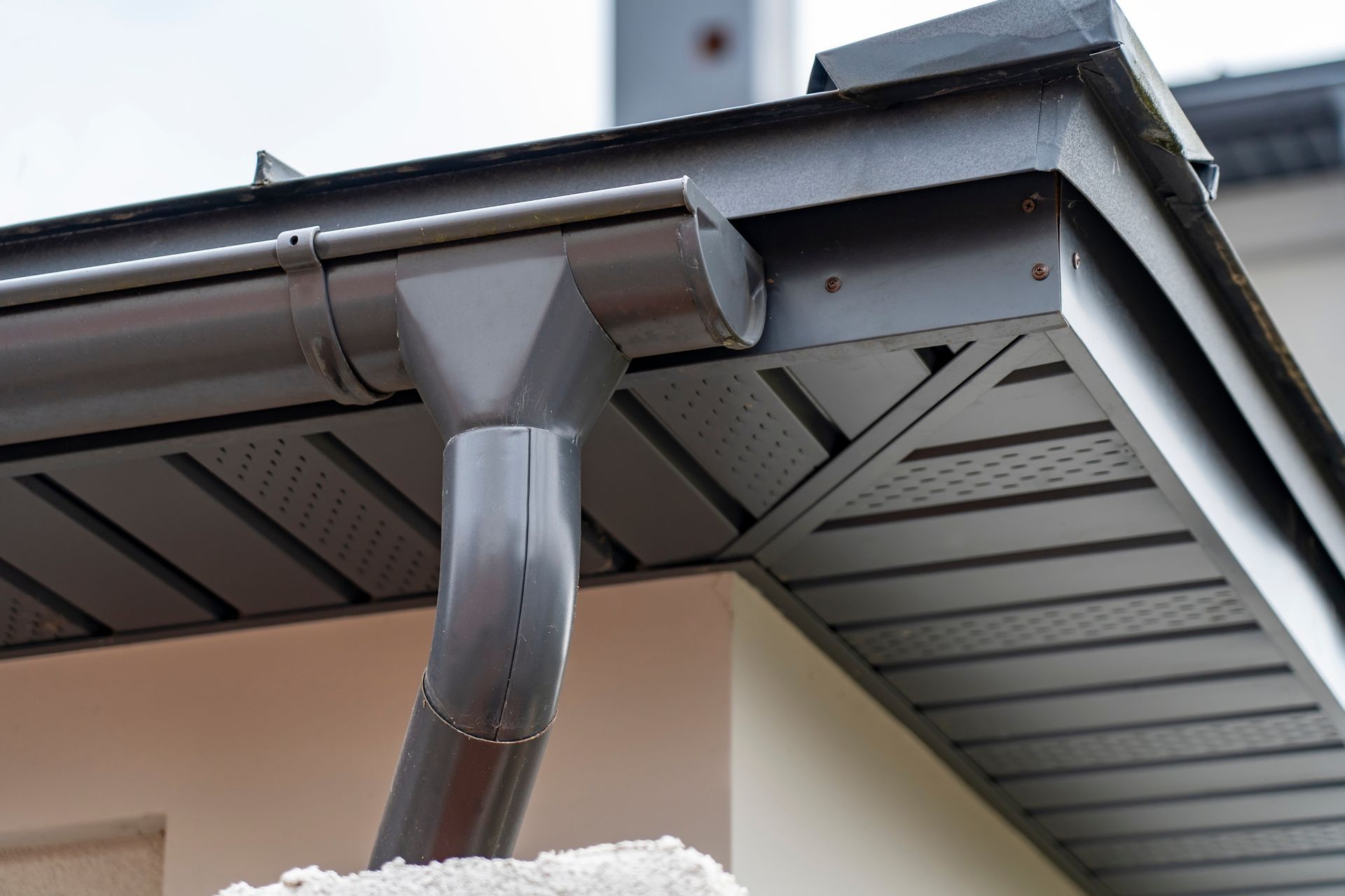 Gray rain gutter system on a house's roof edge. Downspout, fascia, and soffit are visible.