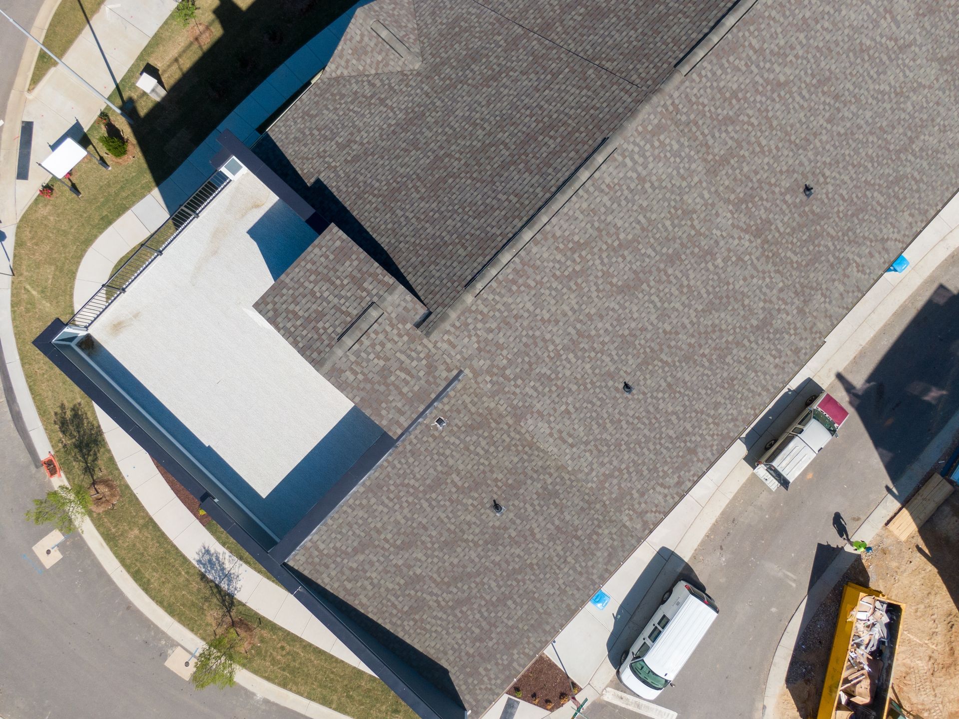 Overhead view of a building with a shingled roof, a white patio, and a curved road.