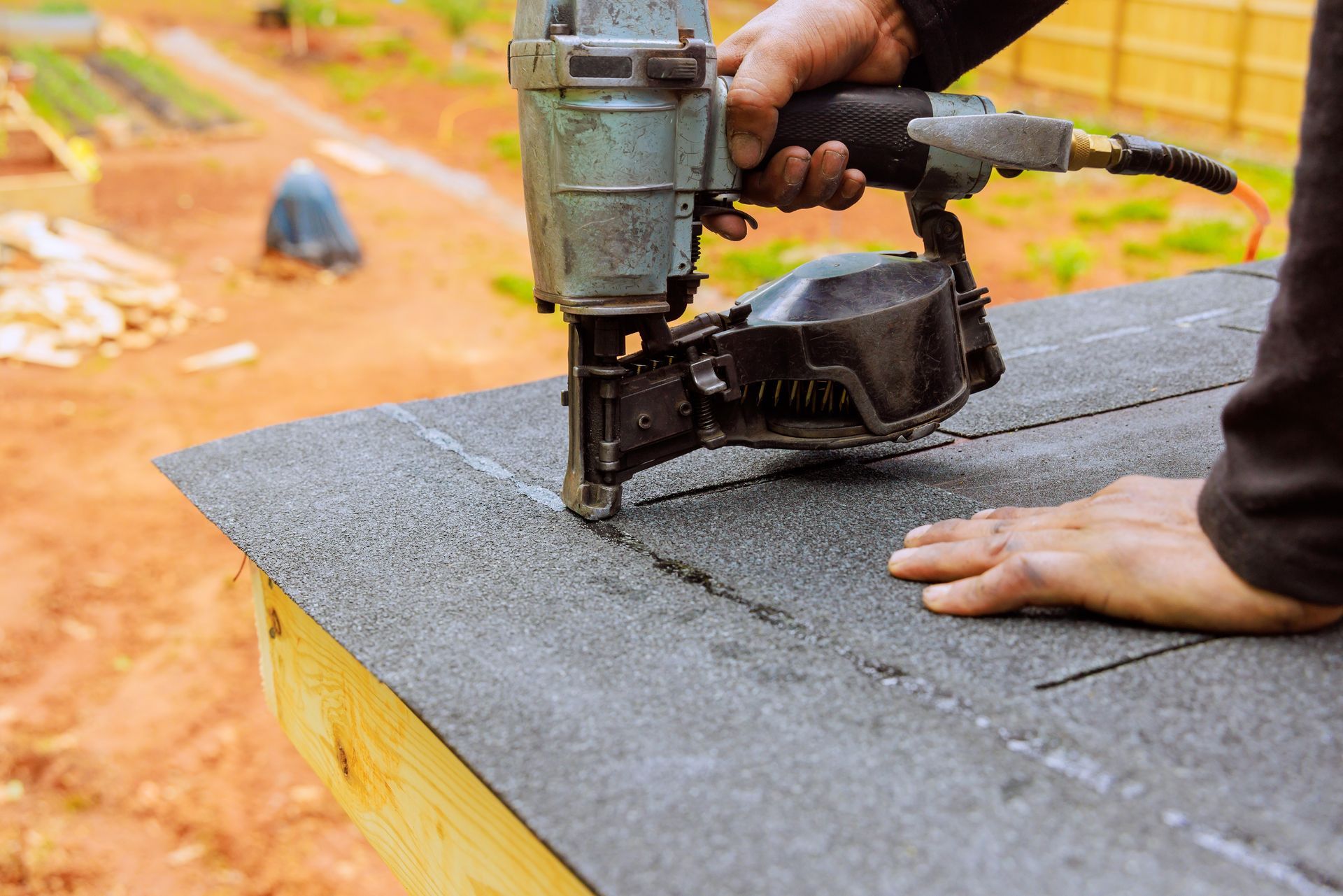 Person using a nail gun to attach dark gray roofing shingles.