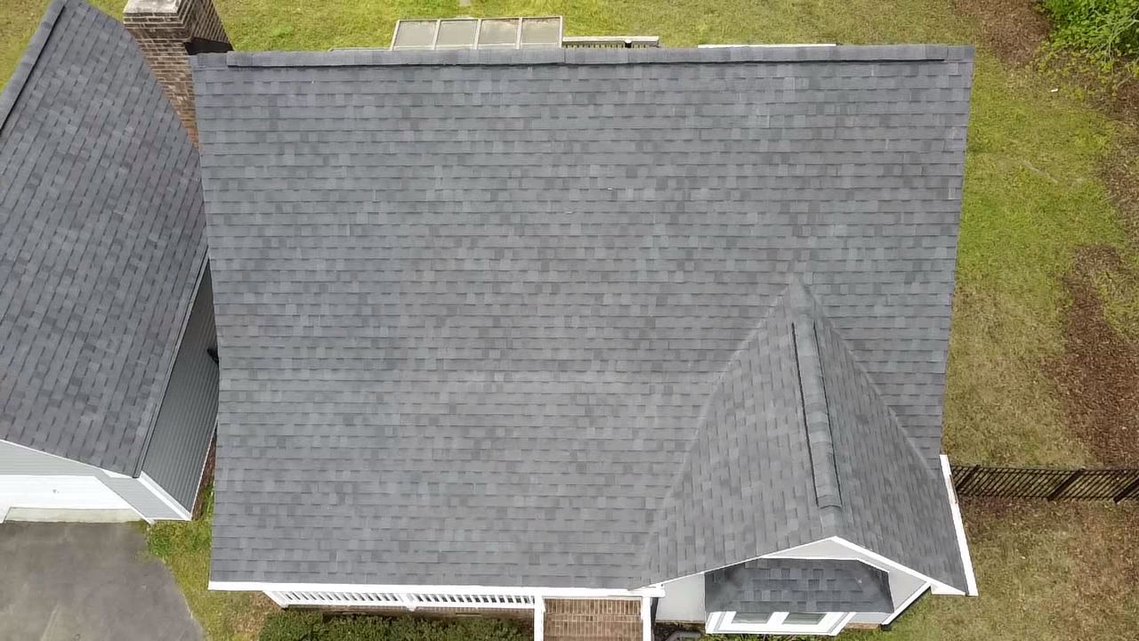 Overhead view of a gray shingled roof with a chimney and part of a house, set on a grassy lawn.