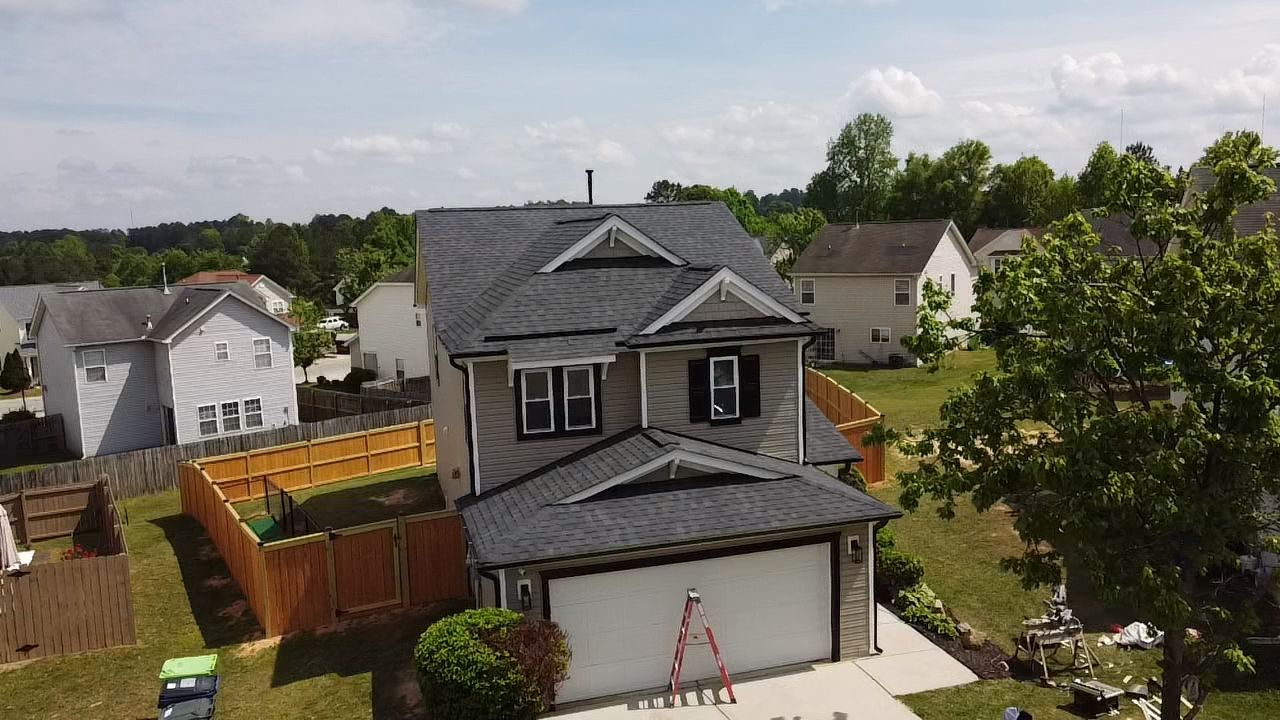 Two-story house with dark gray roof and garage door. A wooden fence surrounds the yard, and a tree stands in the foreground.