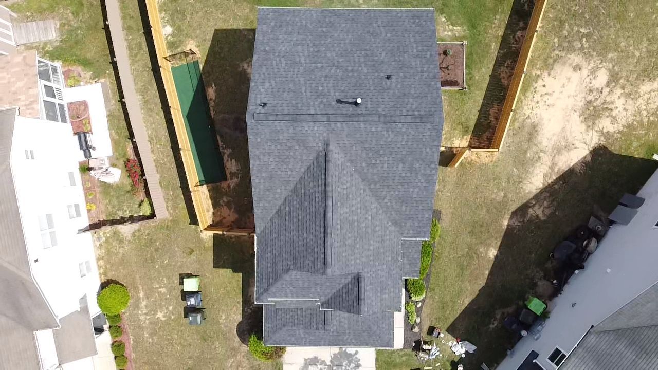 Aerial view of a house with a dark gray roof, surrounded by a yard, trees, and fences.