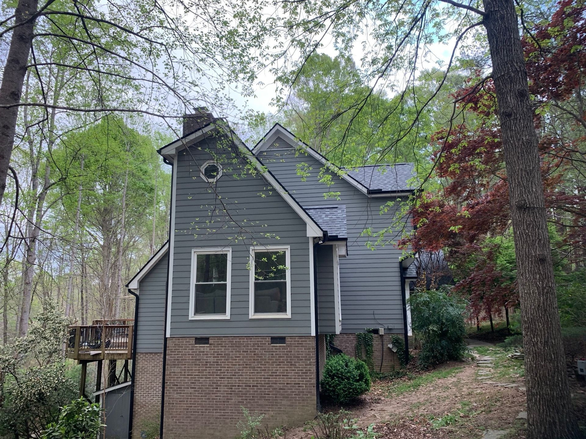 Small gray house with brick base and two windows surrounded by trees.